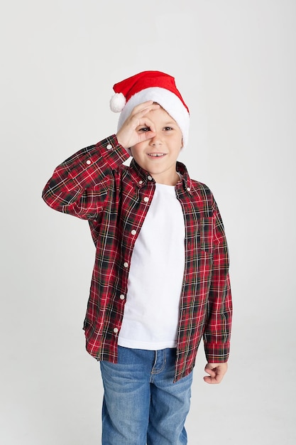 Chłopiec w czerwonej koszulce i czapce Świętego Mikołaja się uśmiecha Śmieszne dziecko w studio na Boże Narodzenie na białym tle Europejskie dziecko uśmiecha się i robi minę Nowy Rok