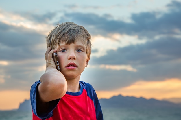 Chłopiec w czerwonej koszulce siedzi na zewnątrz i rozmawia przez telefon komórkowy, wygląda na zdenerwowanego lub przestraszonego
