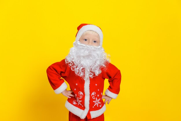 chłopiec w czerwonym stroju Świętego Mikołaja stoi na żółtym tle studia i trzyma ręce na biodrach