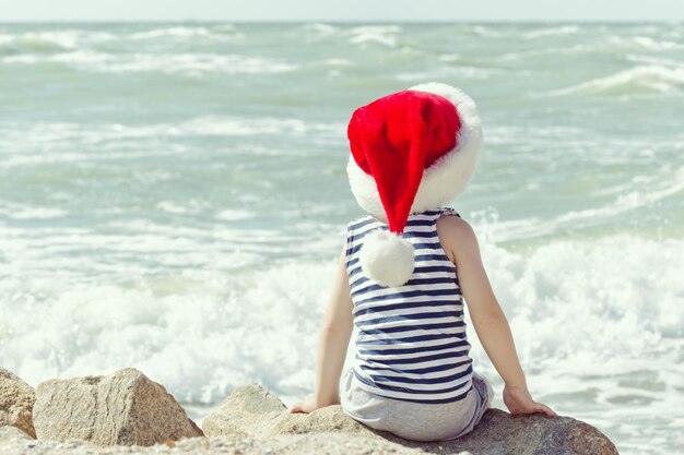 Chłopiec w kapeluszu santa siedzi na skale Brzeg Morza Widok z tyłu