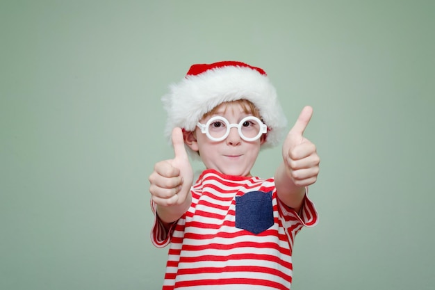 Chłopiec w okularach i kapeluszu Świętego Mikołaja stoi na tle jasnej ściany Fajny gest