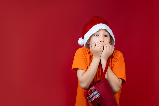 Chłopiec w świątecznym kapeluszu Świętego Mikołaja na czerwonym tle zacisnął pięści przed twarzą ze strachu