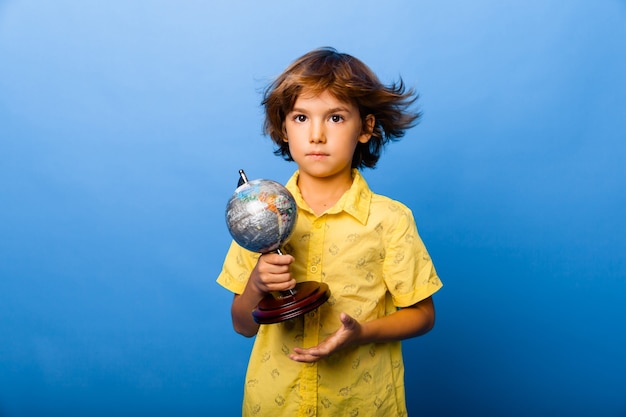 Chłopiec w wieku 7 lat uczeń z pamiątkową kulą ziemską w dłoniach uśmiechający się na niebieskim tle
