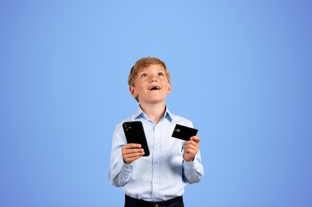 Chłopiec z telefonem i kartą kredytową szczęśliwy portret na pustym błękitnym tle