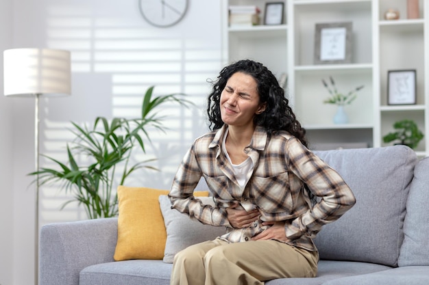 Chora dojrzała dorosła kobieta siedząca samotnie na kanapie w domu z silnym bólem brzucha w Ameryce Łacińskiej
