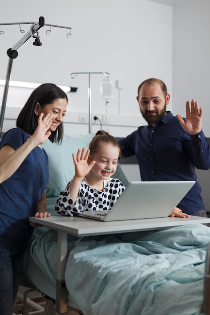 Chora Dziewczynka W Placówce Opieki Zdrowotnej Rozmawia Z Krewnymi Na Wirtualnym Połączeniu Online Na Nowoczesnym Komputerze. Opiekuńczy Rodzice Siedzący Obok Chorego Dziecka Podczas Rozmowy Wideo Na Laptopie.