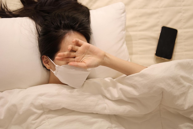 Zdjęcie chorą kobieta, która nosi maskę leżąca w łóżku ręce nad oczami dziewczyna śpiąca w łózku