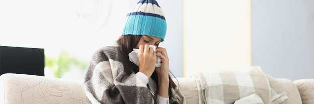 Chora kobieta w ciepłym kapeluszu dmuchająca nos w papierową serwetkę w domu