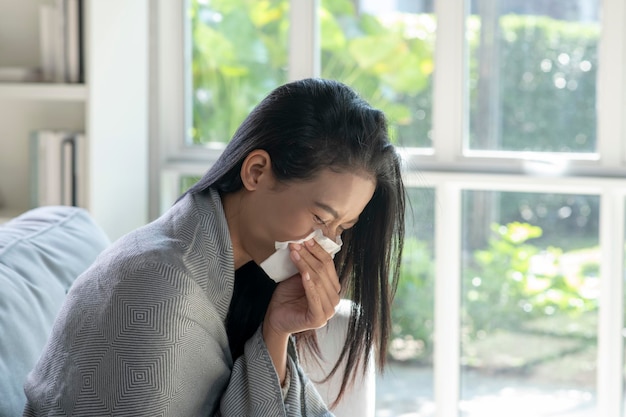 Chora młoda azjatycka kobieta pokryta kocem dmuchającym cieknącym nosem dostała gorączki przeziębienie kichanie w chusteczkę siedzi na kanapie chora alergiczna dziewczyna z objawami alergii kaszel w domu