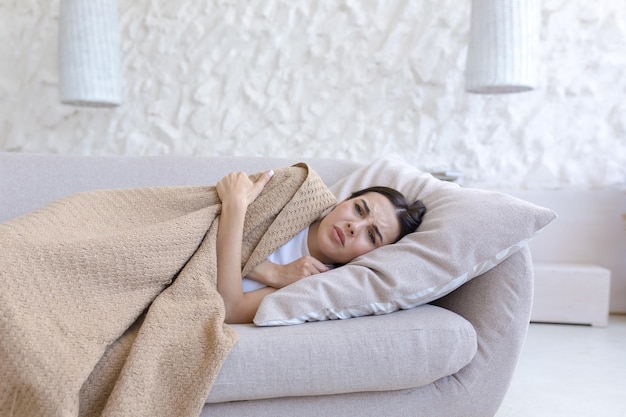 Chora młoda kobieta leżąca na poduszce na kanapie w domu przykryta brązowym kocem czuje się źle