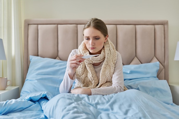 Zdjęcie chora młoda kobieta z termometrem w dłoniach siedzi w domu w łóżku