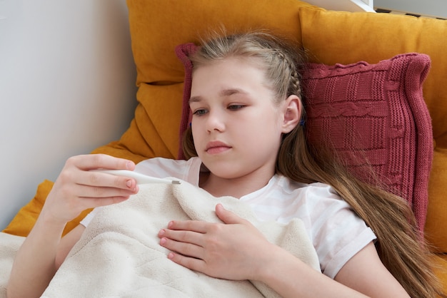 Zdjęcie chora nastoletnia dziewczyna kłaść w łóżku i patrzeje termometr