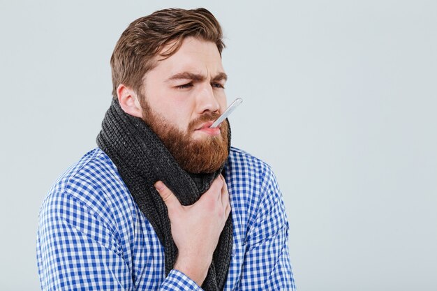 Chory brodaty młody mężczyzna w szaliku trzymający w ustach termometr mierzący temperaturę