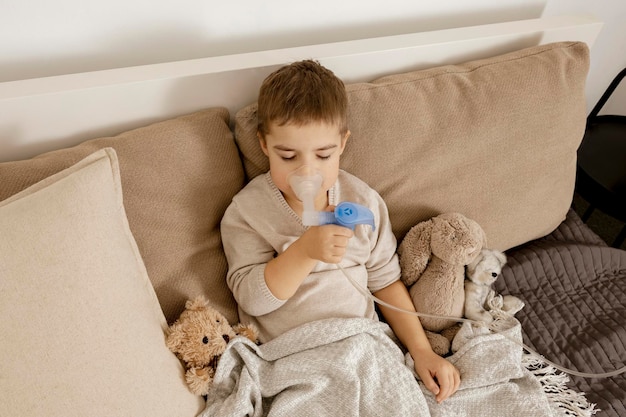 Chory chłopczyk z inhalatorem do leczenia kaszlu. Chory dzieciak robi inhalację na swoim łóżku. Sezon grypowy. Procedura medyczna w domu. Wnętrze i ubrania w naturalnych kolorach ziemi. Przytulne otoczenie.