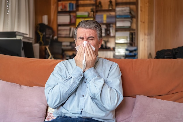 Chory mężczyzna z grypą i kichający do chusteczki siedzący w domu