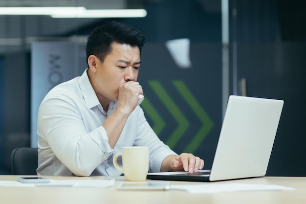 Chory w pracy młody biznesmen z Azji ciągle kaszle zakrywa usta przy biurku w biurze