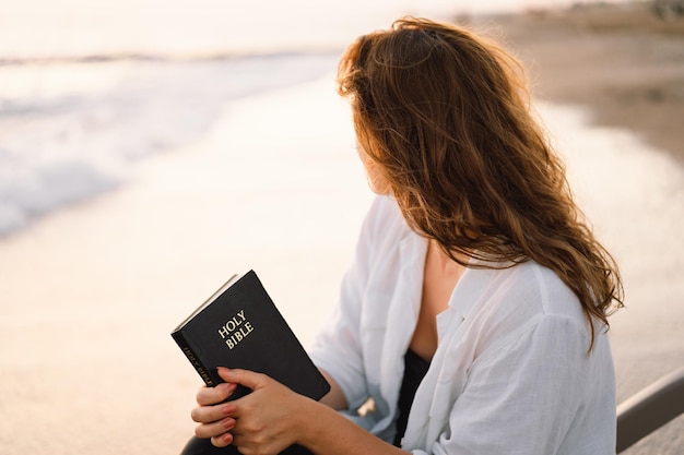 Chrześcijanka trzyma Biblię w dłoniach czytając Pismo Święte nad morzem podczas pięknego zachodu słońca
