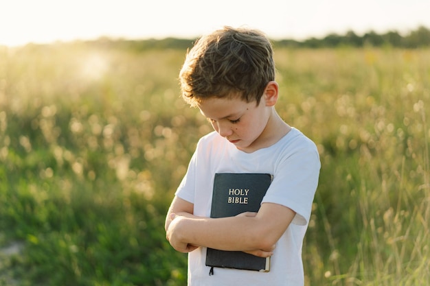 Chrześcijański chłopiec trzyma Biblię w dłoniach Czytanie Pisma Świętego w polu podczas pięknego zachodu słońca Koncepcja wiary duchowości i religii Nadzieja pokoju