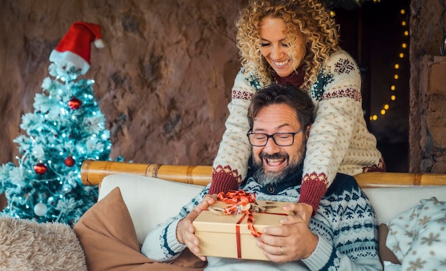 Zdjęcie chwila wymiany prezentów świątecznych w domu z szczęśliwą kobietą dającą mężczyźnie niespodziankę prezent zza kanapy choinka i dekoracje w tle czas wakacji dla zakochanej pary święto zimy
