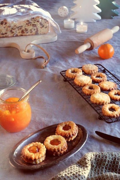 Ciasteczka Yummy Linzer wypełnione dżemem pomarańczowym na stojaku chłodzącym i metalowej płycie w stylu vintage. Boże Narodzenie stollen na stole z naturalnym lnianym obrusem. Skradzione Boże Narodzenie. Naturalne światło słoneczne z długimi cieniami.