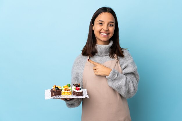 Ciasto szef kuchni trzyma duży tort na białym tle niebieski, wskazując na bok, aby przedstawić produkt.