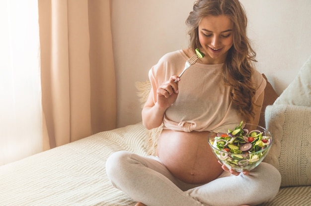 Ciąża i zdrowe odżywianie organiczne. Kobieta w ciąży, ciesząc się sałatką ze świeżych warzyw w łóżku, wolne miejsce