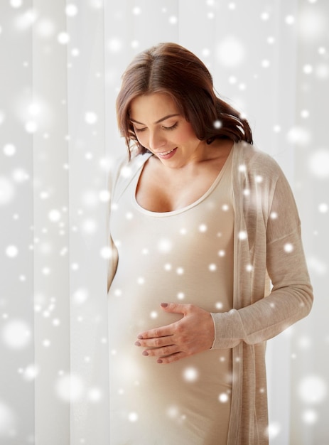 ciąża, macierzyństwo, ludzie, zima i koncepcja oczekiwań - szczęśliwa kobieta w ciąży z dużym brzuszkiem w domu nad śniegiem