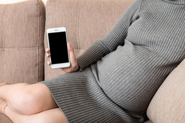 Ciąża z nowoczesnymi technologiamiKobieta w ciąży pokazująca telefon komórkowy z pustym wyświetlaczem na kanapie w domu