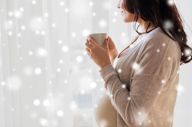 ciąża, zima, święta, ludzie i koncepcja oczekiwań - zbliżenie szczęśliwej kobiety w ciąży z filiżanką picia herbaty patrzącej przez okno w domu na śniegu