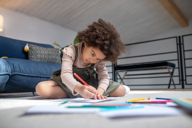 Ciekawa aktywność. Poważna skoncentrowana mała afroamerykańska dziewczynka rysująca kredkami na kartce papieru siedząca na podłodze w pokoju