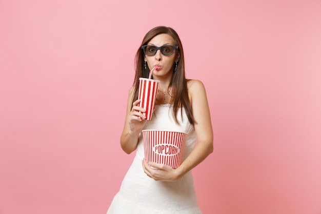 Ciekawa kobieta w białej sukni, okularach 3d oglądająca film, trzymająca wiadro popcornu, plastikowy kubek napoju gazowanego lub coli