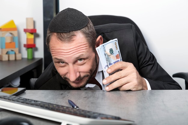 Zdjęcie ciekawie wyglądający mężczyzna brodaty żyd w jarmułce siedzi przy biurku i patrzy na kamerę trzymając studolarowe banknoty szczęśliwy podekscytowany izraelczyk o przebiegłych oczach w biurze wygląda spod stołu