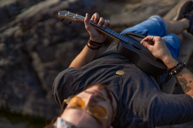 Ciekawy hipis grający na czarnym ukulele leżący na drewnianym moście między skałami o wysokiej k...