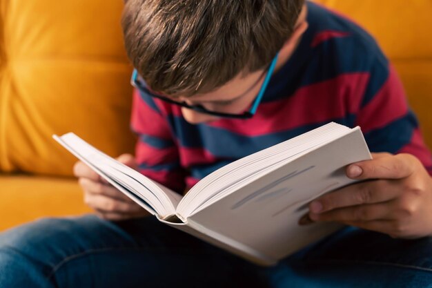 Ciekawy młody chłopiec w okularach jest pochłonięty książką, siedząc wygodnie na kanapie w domu.