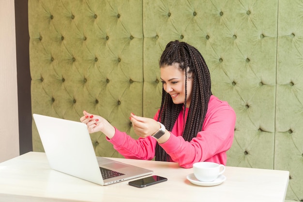 Zdjęcie ciekawy młody freelancer z czarnymi dredami uczesanymi w różową bluzkę siedzi w kawiarni i zgadza się ze swoim pracodawcą na zapłatę przez kamerkę internetową i pokazując gest gotówki z uśmiechniętą buzią. wnętrz