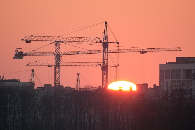 Ciemna sylwetka żurawi wieżowych z dużym zachodzącym słońcem na wysokim placu budowy budynków mieszkalnych o zachodzie słońca