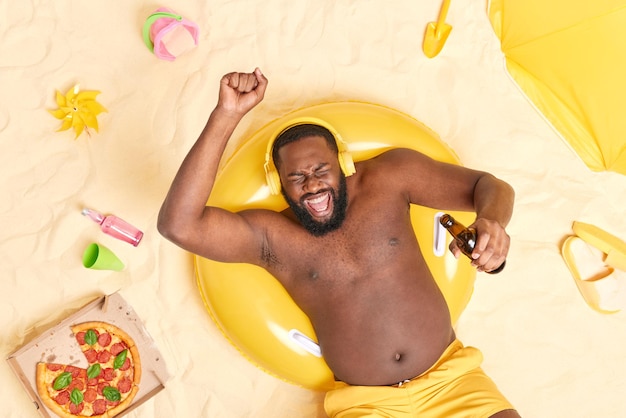 Ciemnoskóry brodaty mężczyzna z nadwagą lubi ulubioną muzykę, potrząsa rękami, pije piwo pozuje na napompowanym basenie na plaży, ma nagi tors otoczony różnymi przedmiotami na piasku Dobra koncepcja odpoczynku