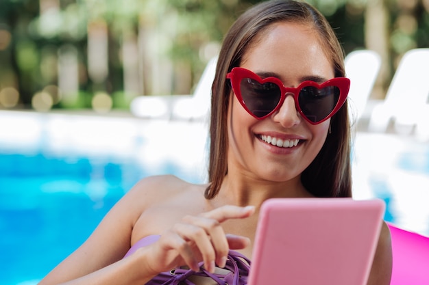 Ciemnowłosa Kobieta śmiejąca Się Noszenie Jasnych Czerwonych Okularów Przeciwsłonecznych, Czytanie Wiadomości Na Swoim Laptopie