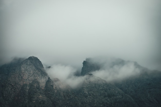 Ciemny atmosferyczny górski krajobraz z ostrymi skałami w niskich chmurach. Ciemne skały w szarą pochmurną pogodę. Ponura górska sceneria z szarymi, niskimi chmurami na szorstkich turniach. Minimalizm z górami skalistymi.