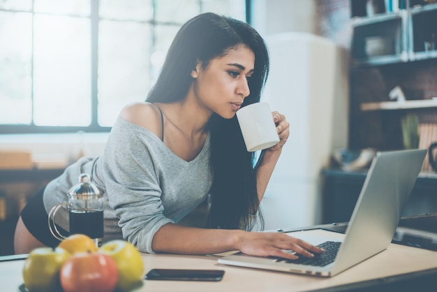 Ciesz się kawą w domu. Piękna młoda kobieta rasy mieszanej pijąca poranną kawę i patrząca na laptopa, opierając się przy biurku w kuchni w domu