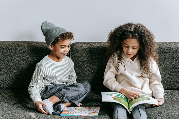 Cieszące się etniczne małe rodzeństwo czytające interesującą książkę na kanapie Zdjęcie