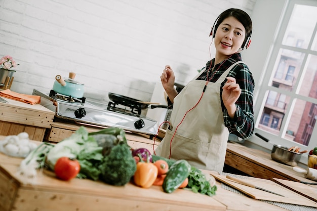 Cieszę się jej porankiem. Piękna młoda azjatycka Japonka w słuchawkach gotowanie sałatki i taniec stojąc w kuchni w domu. szczęśliwa gospodyni domowa w fartuchu poruszająca się z melodią przez słuchawki