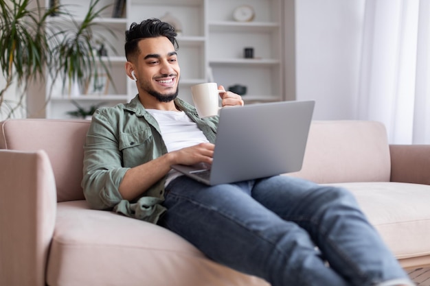 Cieszę się, że milenijny islamski facet z brodą siedzi na kanapie w bezprzewodowych słuchawkach, ogląda laptopa, pije kawę