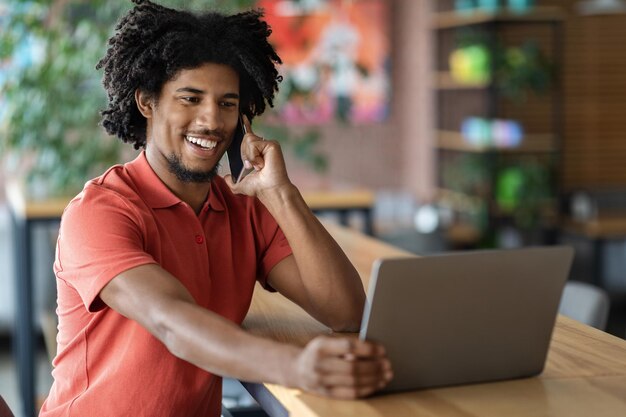 Cieszę się, że młody afroamerykanin kręcony mężczyzna dzwoni przez telefon, patrząc na laptopa przy stole we wnętrzu kawiarni