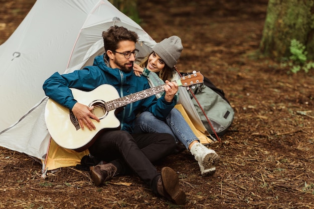 Cieszę się, że tysiącletnia kaukaska kobieta i mężczyzna w kurtkach odpoczywających w zimnym lesie cieszą się przygodą grając na gitarze w pobliżu namiotu