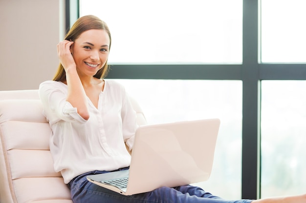 Cieszy się wolnym czasem w domu. Atrakcyjna młoda kobieta pracuje na laptopie i uśmiecha się siedząc na krześle w domu