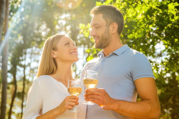 Cieszymy się, że jesteśmy razem. Niski kąt widzenia szczęśliwej młodej pary kochającej trzymającej szklanki z białym winem i patrzącej na siebie z uśmiechem, stojąc razem na zewnątrz