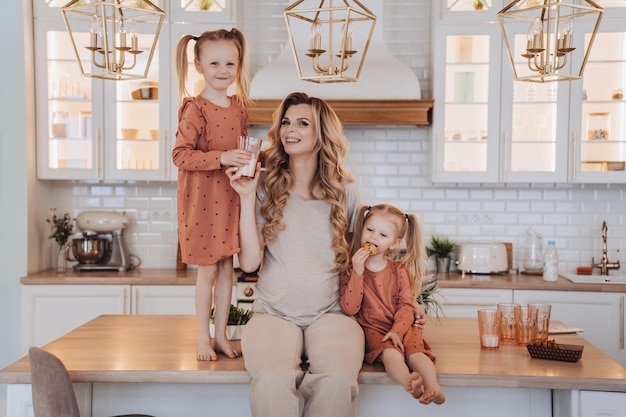 Ciężarna szwedzka mama siedzi na kuchennym stole z małymi córkami ubranymi w beżowe ubrania codzienne