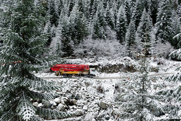 Ciężarówka z dźwigiem jedzie górską drogą, pośród zaśnieżonych wysokich świerków.