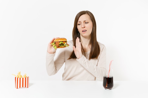 Ciężka kobieta pokazuje gest zatrzymania przy stole z burger frytki cola w szklanej butelce na białym tle. Prawidłowe odżywianie lub amerykańskie klasyczne fast foody. Powierzchnia reklamowa z miejscem na kopię.